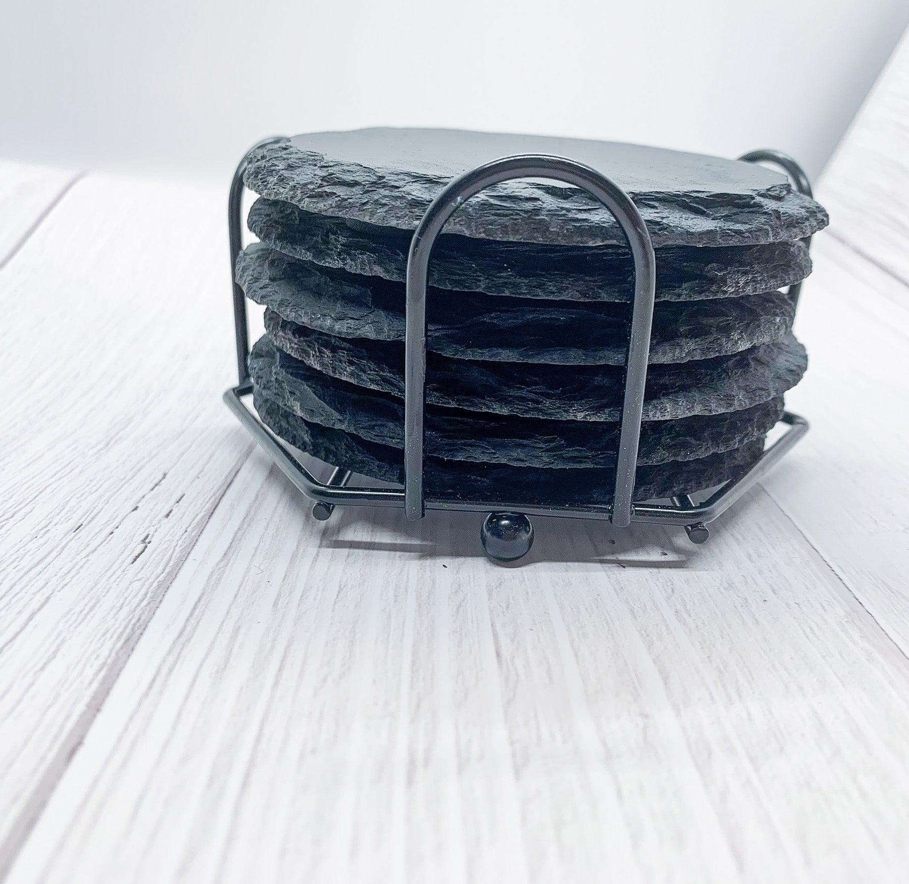 Stack of dark gray natural slate coasters stored in a matching black metal cradle holder on a light wood surface.